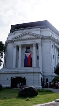 A grand, white neoclassical building with tall columns and a triangular pediment. A large flag is draped between two of the columns. People can be seen gathered at the entrance, which is framed by an archway. The structure is surrounded by a well-maintained lawn with trees and a notable rock in the foreground.