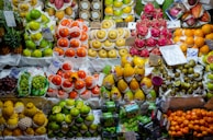A vibrant market stall displays an array of colorful fruits stacked in neat arrangements. The assortment includes oranges, apples, dragon fruits, papayas, grapes, kiwis, and various citrus fruits. Each type of fruit is grouped together, creating a visually appealing and organized presentation.