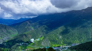 A lush, green mountainous landscape with dense forests stretching across the hills. Buildings, possibly residential or hotels, are nestled within the valleys. The sky above is filled with thick, dramatic clouds casting shadows over the landscape.