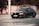 A black sedan car with a license plate number 7P9 8627 is parked on a road. The background includes a red and white striped barrier and dense foliage.