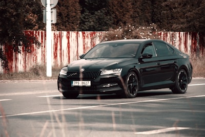 A black sedan car with a license plate number 7P9 8627 is parked on a road. The background includes a red and white striped barrier and dense foliage.