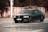 A black sedan car with a license plate number 7P9 8627 is parked on a road. The background includes a red and white striped barrier and dense foliage.