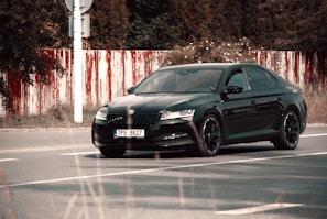 A black sedan car with a license plate number 7P9 8627 is parked on a road. The background includes a red and white striped barrier and dense foliage.
