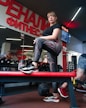 A woman stands confidently on a bench in a gym, dressed in patterned leggings and a black shirt. There is gym equipment such as dumbbells and a medicine ball around her. The interior features a large wall mirror and bright red signage with Cyrillic text. Other gym-goers and gym equipment are visible in the background.
