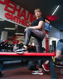 A woman stands confidently on a bench in a gym, dressed in patterned leggings and a black shirt. There is gym equipment such as dumbbells and a medicine ball around her. The interior features a large wall mirror and bright red signage with Cyrillic text. Other gym-goers and gym equipment are visible in the background.