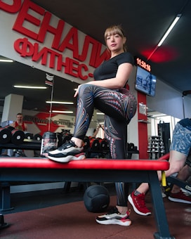 A woman stands confidently on a bench in a gym, dressed in patterned leggings and a black shirt. There is gym equipment such as dumbbells and a medicine ball around her. The interior features a large wall mirror and bright red signage with Cyrillic text. Other gym-goers and gym equipment are visible in the background.