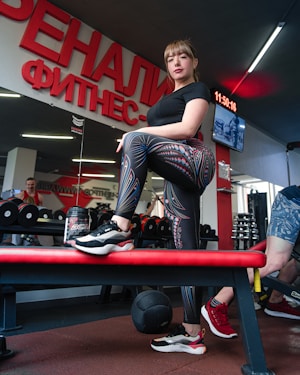 A woman stands confidently on a bench in a gym, dressed in patterned leggings and a black shirt. There is gym equipment such as dumbbells and a medicine ball around her. The interior features a large wall mirror and bright red signage with Cyrillic text. Other gym-goers and gym equipment are visible in the background.