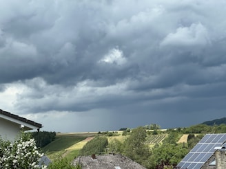 Dark storm clouds hover over a rural landscape with green fields and patches of forest. In the foreground, rooftops with solar panels and flowering bushes are visible.