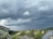 Dark storm clouds hover over a rural landscape with green fields and patches of forest. In the foreground, rooftops with solar panels and flowering bushes are visible.