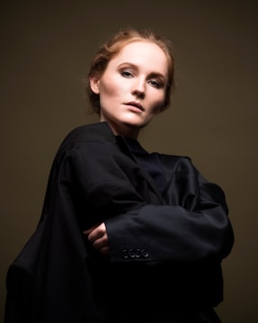 A person gazes intently at the camera, wearing a dark, oversized coat that wraps around their shoulders. Their hair is styled neatly, and the backdrop is a subdued, muted tone that highlights their facial features.