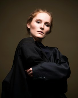 A person gazes intently at the camera, wearing a dark, oversized coat that wraps around their shoulders. Their hair is styled neatly, and the backdrop is a subdued, muted tone that highlights their facial features.