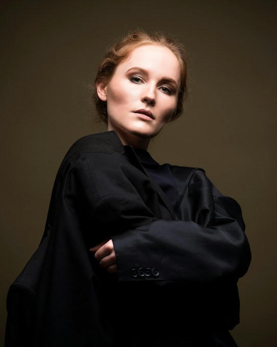A person gazes intently at the camera, wearing a dark, oversized coat that wraps around their shoulders. Their hair is styled neatly, and the backdrop is a subdued, muted tone that highlights their facial features.
