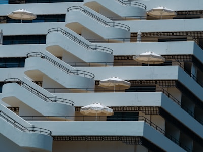 An architectural design featuring multiple stacked balconies with black railings and white parasols. The balconies are arranged in a cascading pattern, creating a visual rhythm across the building facade.