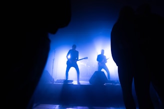 A band is performing on stage with a silhouette effect caused by bright blue lighting. The musicians are playing guitars, and the stage setup includes microphones and speakers. The audience appears as dark silhouettes framing the image.
