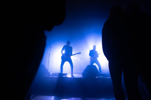 A band is performing on stage with a silhouette effect caused by bright blue lighting. The musicians are playing guitars, and the stage setup includes microphones and speakers. The audience appears as dark silhouettes framing the image.