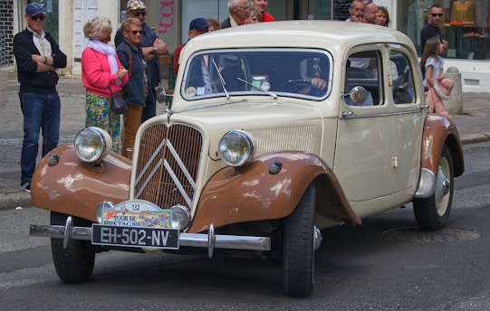 An antique cream and brown car is on a street lined with a group of onlookers. The vehicle, featuring vintage styling, is adorned with a license plate reading 'EH-502-NV' and a front badge that appears related to a car rally or event. The audience includes people of various ages, dressed casually, some standing on the sidewalk, appearing interested in the car.