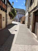 A narrow cobblestone street in a charming village with half-timbered houses and buildings. The street is flanked by quaint, old buildings with wooden shutters. A vintage street lamp hangs on the left side, and there is bright sunlight casting shadows along the street.