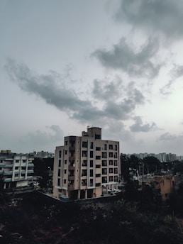 A multi-story residential building stands prominently in an urban setting under a cloudy sky. The building features modern architectural design with balconies and is surrounded by other structures and greenery. The sky is overcast with thick, gray clouds adding a dramatic effect.
