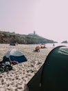 A sandy beach with several tents set up along the shoreline. People are lounging around, enjoying the day near the ocean. In the background, a rocky cliff supports a lighthouse overlooking the sea.