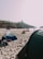 A sandy beach with several tents set up along the shoreline. People are lounging around, enjoying the day near the ocean. In the background, a rocky cliff supports a lighthouse overlooking the sea.