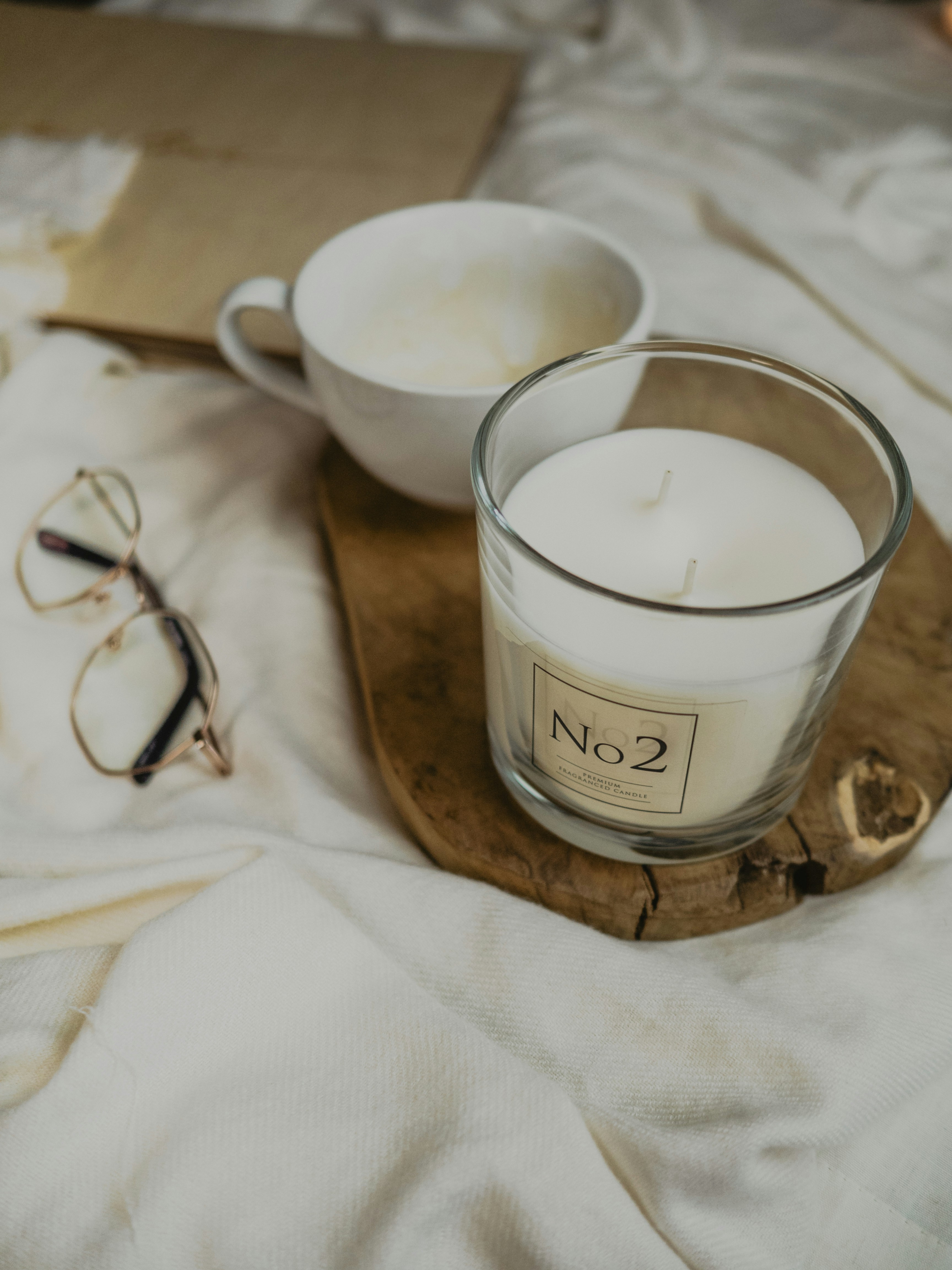 A coffee cup and candle on a wooden board, next to a pair of glasses, all on a white cloth