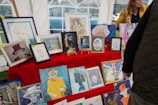A collection of framed artworks displayed on a red cloth-covered table, mostly featuring portraits. The artworks vary in style, including sketches and painted portraits. A woman in a yellow jacket stands nearby, possibly discussing the artworks.