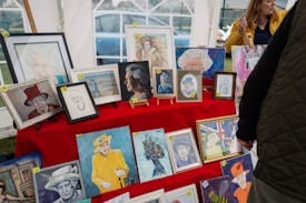 A collection of framed artworks displayed on a red cloth-covered table, mostly featuring portraits. The artworks vary in style, including sketches and painted portraits. A woman in a yellow jacket stands nearby, possibly discussing the artworks.
