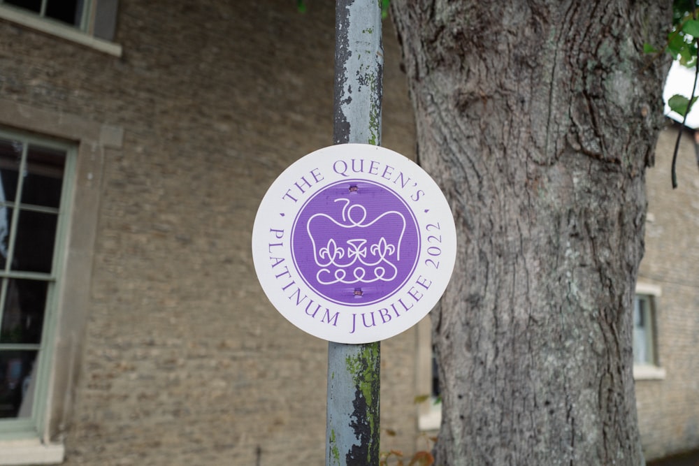 a purple and white sign on a pole next to a tree