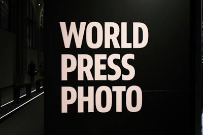 Large bold white letters read 'WORLD PRESS PHOTO' against a dark background. The setting is likely an exhibition or gallery with dim lighting and architectural features visible at the back.