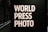 Large bold white letters read 'WORLD PRESS PHOTO' against a dark background. The setting is likely an exhibition or gallery with dim lighting and architectural features visible at the back.