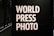Large bold white letters read 'WORLD PRESS PHOTO' against a dark background. The setting is likely an exhibition or gallery with dim lighting and architectural features visible at the back.