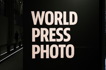 Large bold white letters read 'WORLD PRESS PHOTO' against a dark background. The setting is likely an exhibition or gallery with dim lighting and architectural features visible at the back.