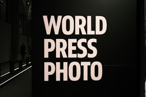 Large bold white letters read 'WORLD PRESS PHOTO' against a dark background. The setting is likely an exhibition or gallery with dim lighting and architectural features visible at the back.