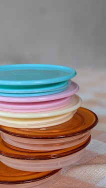A stack of round, plastic lids in various colors including blue, pink, cream, and amber is neatly arranged on a surface. The lids are translucent and each layer is visible through the others, creating a subtle blending of shades. The background is softly blurred, emphasizing the focus on the lids.
