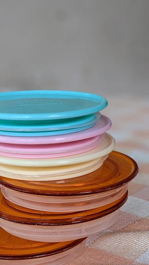 A stack of round, plastic lids in various colors including blue, pink, cream, and amber is neatly arranged on a surface. The lids are translucent and each layer is visible through the others, creating a subtle blending of shades. The background is softly blurred, emphasizing the focus on the lids.