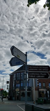 A street scene with directional signs pointing to various locations including Slane and Dublin. The signs are in both English and Irish. Behind the signs, there is a building with a round corner and large windows, housing businesses like a public library and a place named Royal. The sky is filled with scattered clouds.