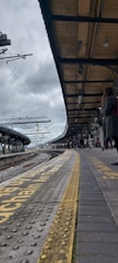 A train platform under a cloudy sky, with overhead electric lines and a wooden roof structure. The perspective focuses on the tracks leading into the distance. There are several people waiting along the platform, some standing and a few sitting. The platform edge is marked with textured yellow warning lines and signage.