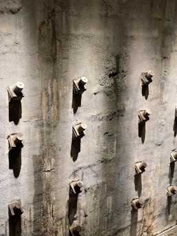 A textured concrete wall with an array of evenly spaced, protruding bolts. The surface appears weathered with some rust stains around the bolts, and it catches angled sunlight, forming shadows that add depth to the structure.