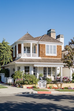 A two-story house with a brown shingle roof and a white exterior. The upper level features a balcony with white railings, while the lower level has large windows and a striped awning. The front yard is well-kept with numerous plants and flowers, and a white picket fence borders the property. The house is situated on a corner lot with a sidewalk and a well-maintained street.
