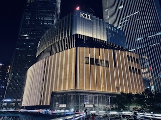 A modern skyscraper at night with prominent vertical lighting patterns. The building's exterior features a combination of dark and illuminated sections, giving it a sophisticated appearance. The name 'K11' is visible at the top. Surrounding structures and some greenery are partially visible.