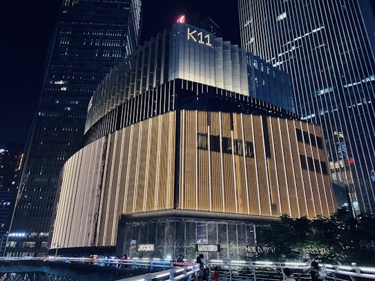 A modern skyscraper at night with prominent vertical lighting patterns. The building's exterior features a combination of dark and illuminated sections, giving it a sophisticated appearance. The name 'K11' is visible at the top. Surrounding structures and some greenery are partially visible.