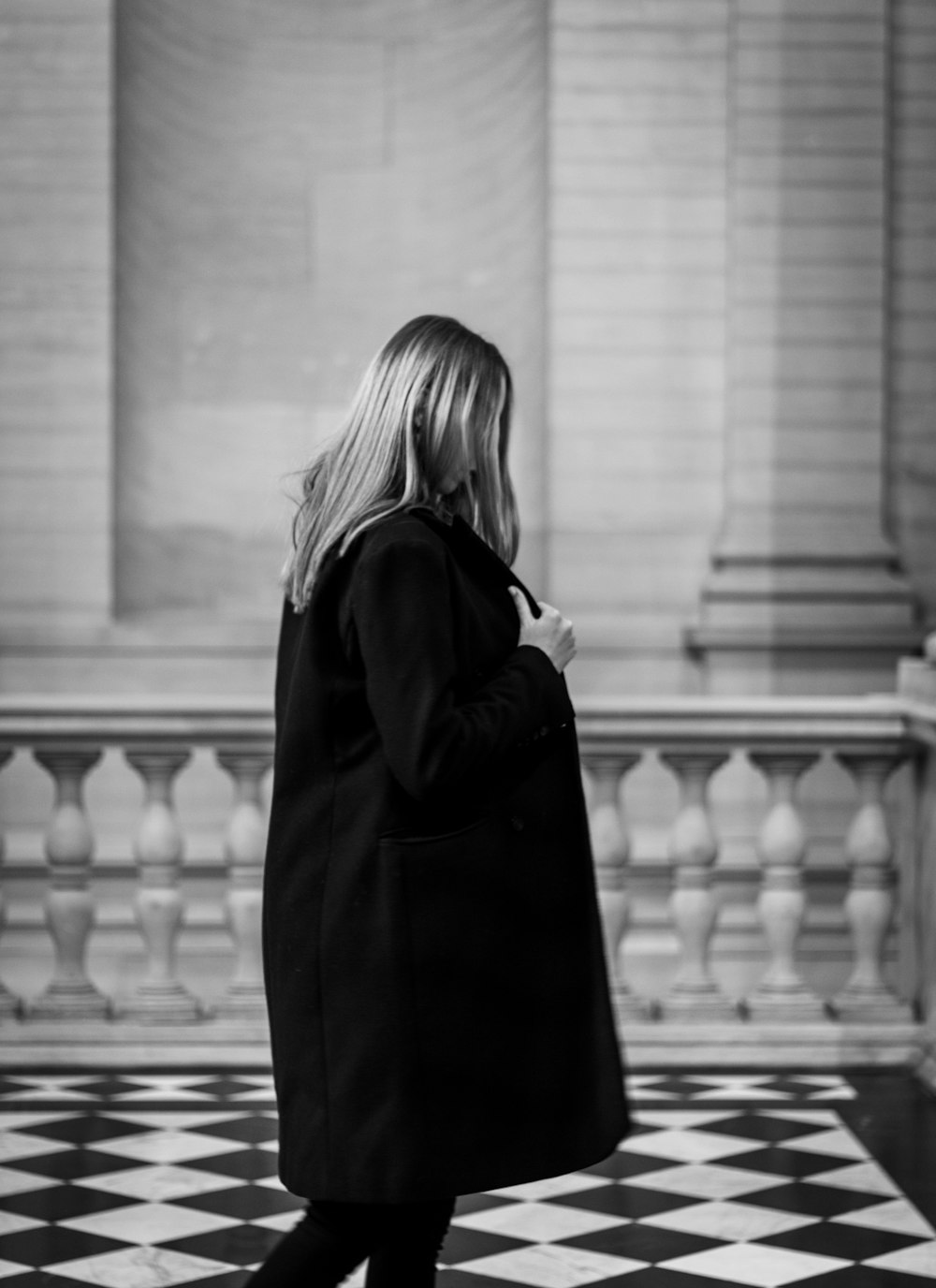 a woman in a black coat is walking down a checkered floor
