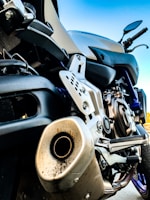 Close-up of a motorcycle exhaust and engine parts, showing the intricate details of the metal components and structure. The image captures the lower section of the motorcycle with a focus on the engine casing, exhaust pipe and footrest. The motorcycle appears to be parked, with handlebars visible in the upper right portion of the image against a clear blue sky background.