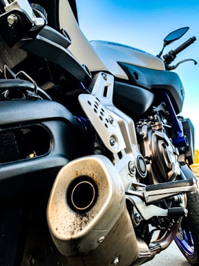 Close-up of a motorcycle exhaust and engine parts, showing the intricate details of the metal components and structure. The image captures the lower section of the motorcycle with a focus on the engine casing, exhaust pipe and footrest. The motorcycle appears to be parked, with handlebars visible in the upper right portion of the image against a clear blue sky background.