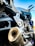 Close-up of a motorcycle exhaust and engine parts, showing the intricate details of the metal components and structure. The image captures the lower section of the motorcycle with a focus on the engine casing, exhaust pipe and footrest. The motorcycle appears to be parked, with handlebars visible in the upper right portion of the image against a clear blue sky background.