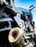 Close-up of a motorcycle exhaust and engine parts, showing the intricate details of the metal components and structure. The image captures the lower section of the motorcycle with a focus on the engine casing, exhaust pipe and footrest. The motorcycle appears to be parked, with handlebars visible in the upper right portion of the image against a clear blue sky background.