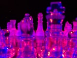 A collection of translucent chess pieces are illuminated by colorful neon lights, casting vibrant hues across a reflective surface. The pieces are arranged on a chessboard, with a striking contrast between the bright blues and pinks against a dark background.