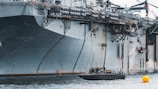A large naval aircraft carrier with visible identification letters is stationed in the water. On the deck, various equipment and a helicopter are present. Below, a smaller patrol boat, manned by military personnel, navigates near a bright yellow buoy. The scene appears industrial and maritime.