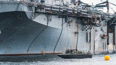 A large naval aircraft carrier with visible identification letters is stationed in the water. On the deck, various equipment and a helicopter are present. Below, a smaller patrol boat, manned by military personnel, navigates near a bright yellow buoy. The scene appears industrial and maritime.