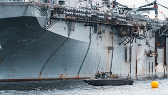 A large naval aircraft carrier with visible identification letters is stationed in the water. On the deck, various equipment and a helicopter are present. Below, a smaller patrol boat, manned by military personnel, navigates near a bright yellow buoy. The scene appears industrial and maritime.
